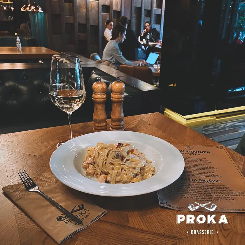 Pjatë me pasta, gotë vere dhe menu e printuar në një tavolinë në Proka
